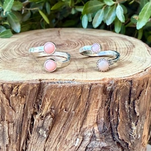 May include: Two open-style silver rings, each featuring two round, light pink cabochon stones. The metal bands have a hammered, textured finish. One ring displays smooth, polished stone settings, while the other ring features scalloped, decorative edges around the pink stones.