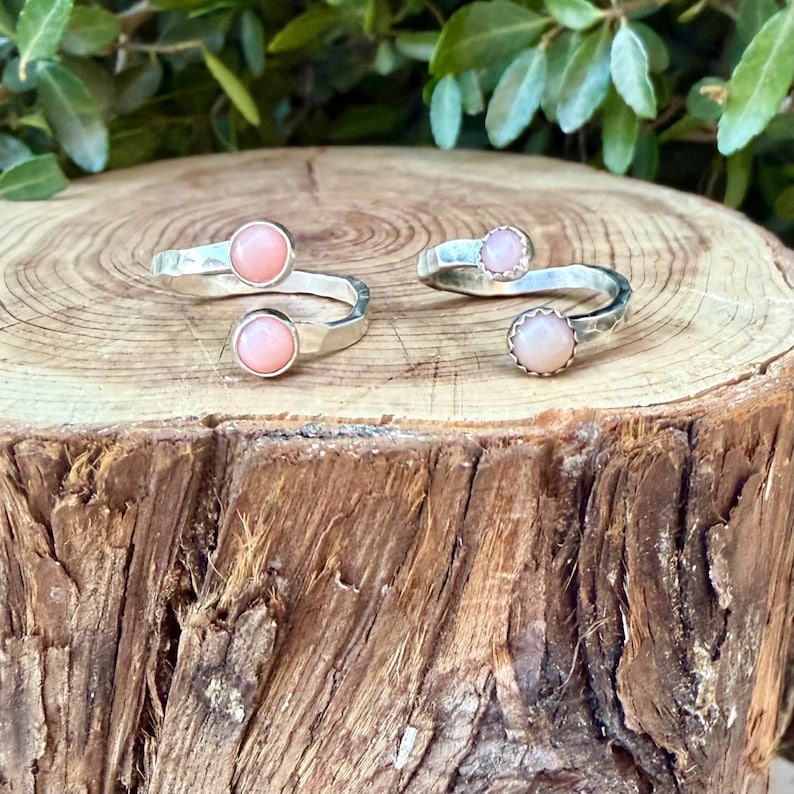 May include: Two open-style silver rings, each featuring two round, light pink cabochon stones. The metal bands have a hammered, textured finish. One ring displays smooth, polished stone settings, while the other ring features scalloped, decorative edges around the pink stones.
