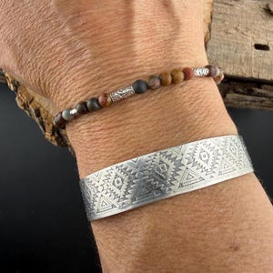 May include: A beaded bracelet featuring small, multi-colored matte stone beads interspersed with silver-toned metal spacers, worn alongside a wide, flat silver-toned metal cuff bracelet engraved with a repeating geometric pattern.