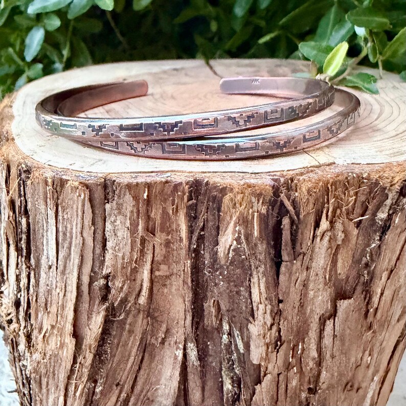 May include: A copper cuff bracelet featuring a repeating geometric pattern etched along the outer surface. The metal has a warm, reddish-brown metallic finish with dark oxidised detailing within the engraved lines of the design.