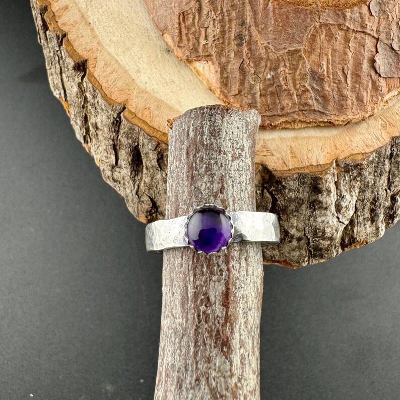 May include: A silver ring featuring a hammered band and a round, deep purple gemstone. The ring is displayed on a piece of wood with a natural bark texture, set against a dark background.