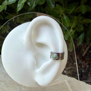 May include: A silver ear cuff with a turquoise stone set in the center. The cuff is detailed with an embossed floral pattern. The ear cuff is shown on a white ear model against a backdrop of green foliage.