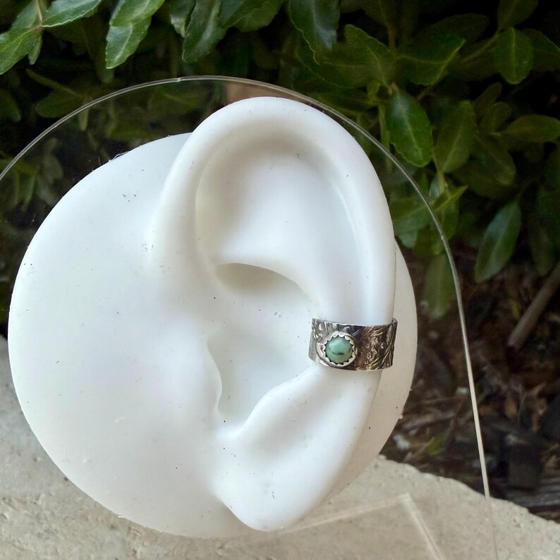 May include: A silver ear cuff with a turquoise stone set in the center. The cuff is detailed with an embossed floral pattern. The ear cuff is shown on a white ear model against a backdrop of green foliage.