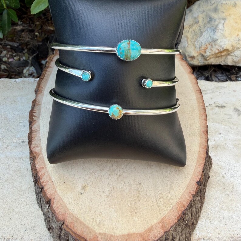 May include: Three silver-toned cuff bracelets with turquoise stones. The bracelets are displayed on a black stand atop a wooden surface. The turquoise stones are oval and round, with a mix of blue and brown hues.