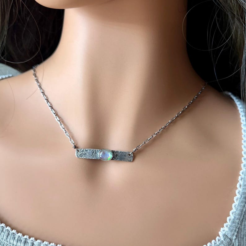 May include: A silver-toned necklace featuring a horizontal rectangular bar pendant with an etched, textured surface. A small, round, iridescent cabochon stone is set in the center of the bar, which hangs from a delicate silver-toned chain.