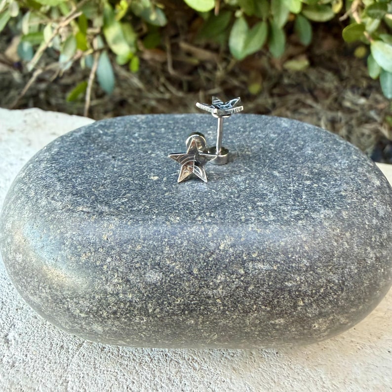 May include: A pair of silver-toned star-shaped stud earrings with a metallic finish. Each earring features a five-pointed star design attached to a straight post backing, showcasing a polished surface that reflects light from multiple angles.