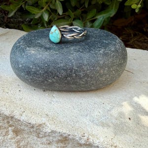 Puede incluir: Un anillo de plata con una piedra turquesa en forma de l&aacute;grima con vetas de matriz marr&oacute;n. La banda del anillo est&aacute; construida con tres hilos de plata entrelazados, creando una textura trenzada que rodea el dedo.