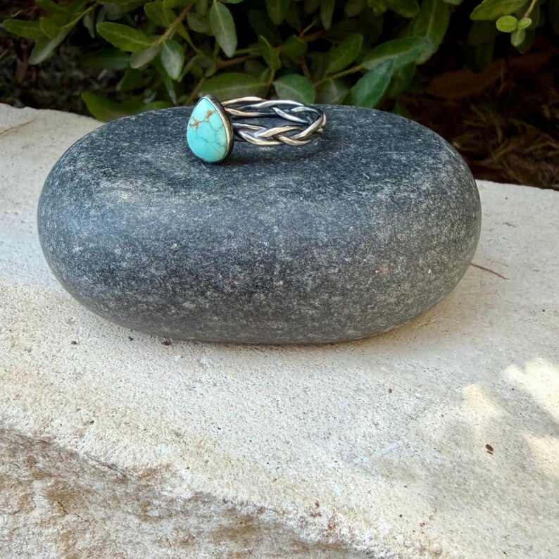 Puede incluir: Un anillo de plata con una piedra turquesa en forma de l&aacute;grima con vetas de matriz marr&oacute;n. La banda del anillo est&aacute; construida con tres hilos de plata entrelazados, creando una textura trenzada que rodea el dedo.