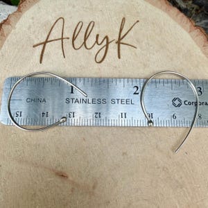 Puede incluir: Dos pendientes de alambre curvado en tono plateado con extremos de peque&ntilde;a bola, colocados sobre una regla de acero inoxidable que muestra un ancho de aproximadamente 6,35 cm. El texto Ally K est&aacute; grabado en la superficie de madera clara.