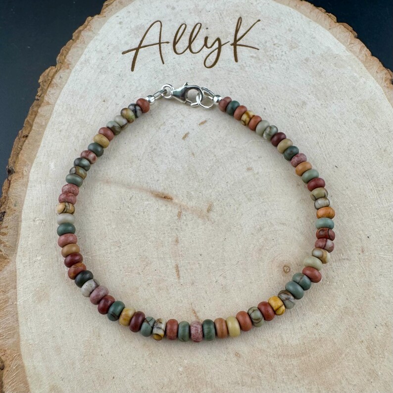 May include: A beaded bracelet with a variety of earth-toned beads, including shades of red, green, brown, and yellow. The bracelet is secured with a silver clasp. The text "Allyk" is written in a script font above the bracelet.