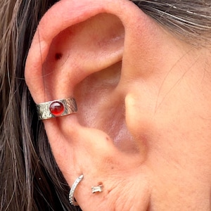 May include: A silver ear cuff with a red gemstone set on the ear. The cuff has a textured design. A small, silver hoop earring and a small, rectangular stud earring are also visible.