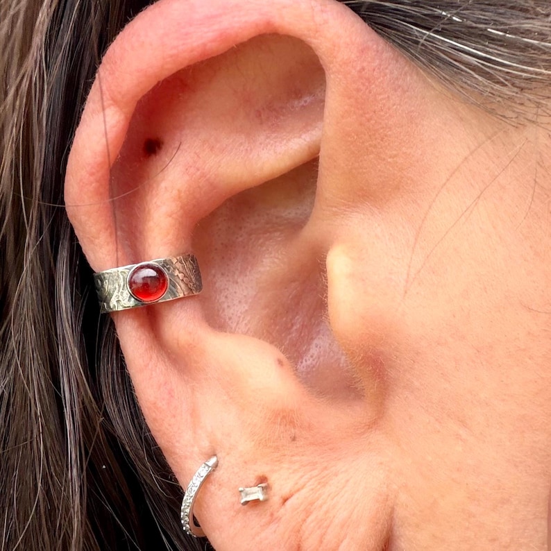 May include: A silver ear cuff with a red gemstone set on the ear. The cuff has a textured design. A small, silver hoop earring and a small, rectangular stud earring are also visible.