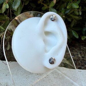 May include: Two small, round, silver-toned stud earrings featuring a stamped cross design with a central dot. The earrings are displayed on a white ear-shaped model stand, showcasing a polished metallic finish and a flat, circular profile.
