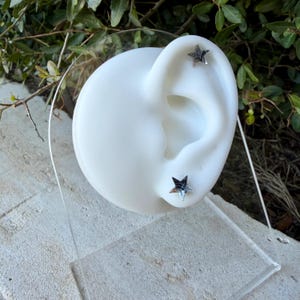 May include: Two small, five-pointed star-shaped stud earrings with a metallic, reflective finish. The earrings are displayed on a white, ear-shaped mannequin stand made of smooth material, positioned against a natural outdoor background.