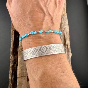 May include: A silver cuff bracelet featuring a repeating geometric pattern of triangles and diamonds, worn alongside a delicate beaded bracelet made of small, bright blue turquoise-colored stones and thin silver spacers.