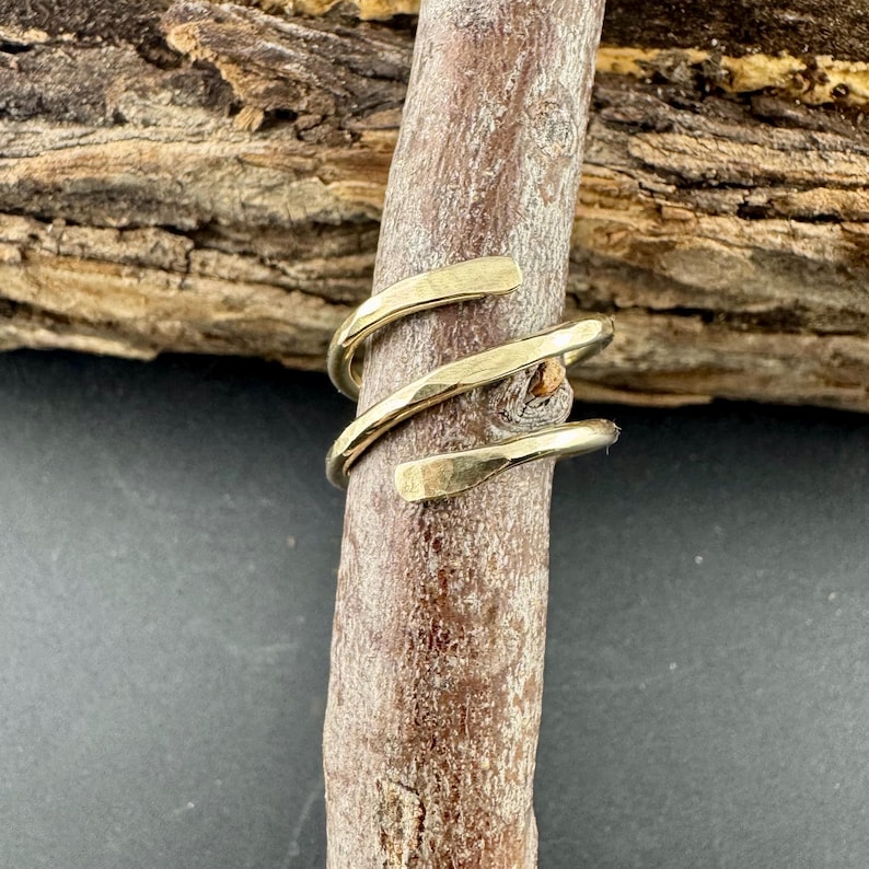 May include: A gold-colored, spiral ring with a hammered texture. The ring wraps around a brown wooden stick, showcasing its design. The ring's design is simple and elegant, suitable for everyday wear.