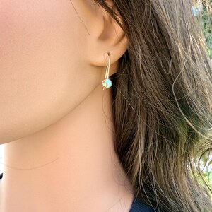 May include: A gold-toned earring featuring a single, small, round turquoise-colored bead with subtle brown veining. The bead is suspended from a thin, curved gold-toned wire hook that passes through the earlobe, creating a simple drop design.