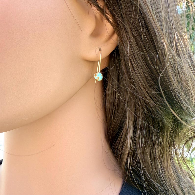 May include: A gold-toned earring featuring a single, small, round turquoise-colored bead with subtle brown veining. The bead is suspended from a thin, curved gold-toned wire hook that passes through the earlobe, creating a simple drop design.