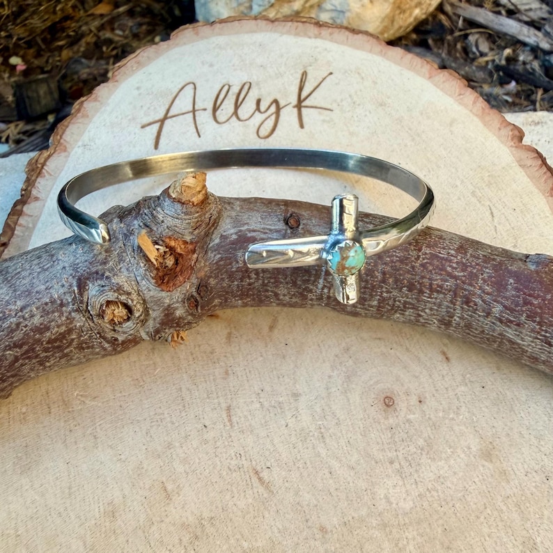 May include: A silver-toned cuff bracelet with a turquoise stone set in a cross design. The bracelet is displayed on a wooden branch, with the name "Ally K" written on a wooden background.