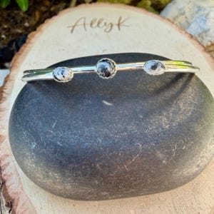 May include: A silver bracelet with three round, speckled stones. The stones are set in a simple, elegant design. The bracelet is displayed on a dark gray stone, with the name "Allyk" lightly etched on a wooden surface in the background.