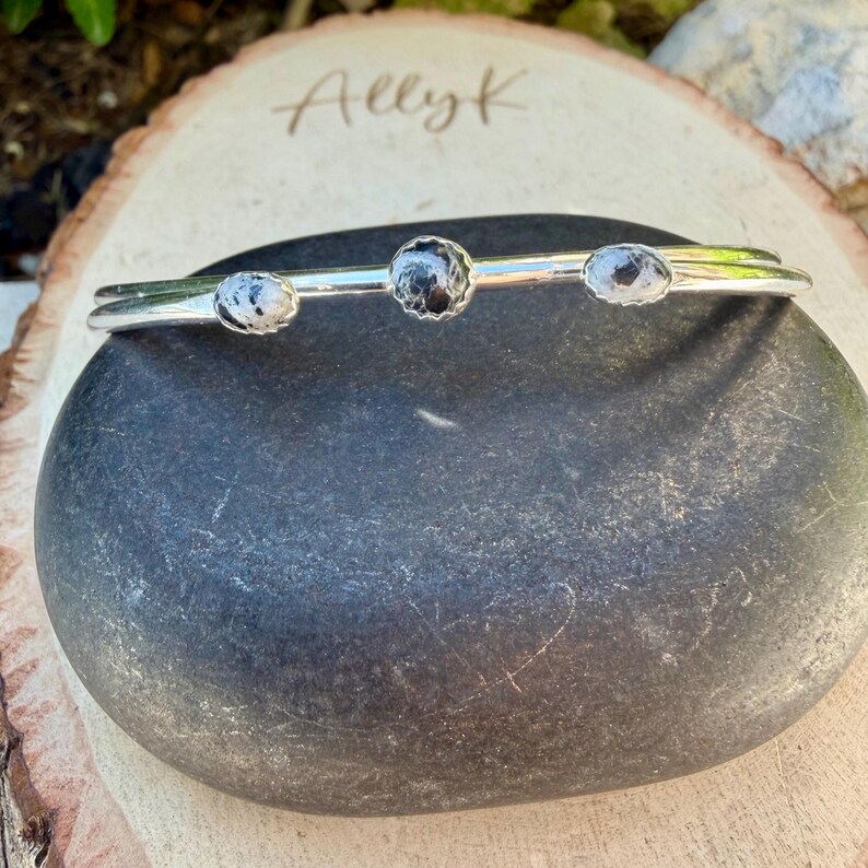 May include: A silver bracelet with three round, speckled stones. The stones are set in a simple, elegant design. The bracelet is displayed on a dark gray stone, with the name "Allyk" lightly etched on a wooden surface in the background.