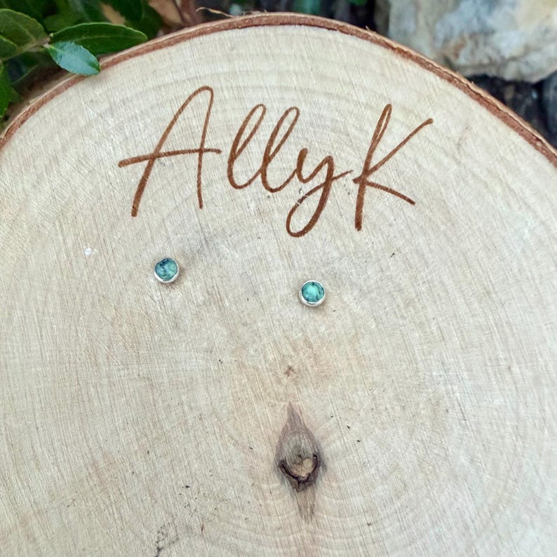 Pu&ograve; includere: Un paio di piccoli orecchini a bottone rotondi con pietre color teal incastonate in argento. Gli orecchini sono esposti su una superficie di legno con il nome "Allyk" inciso in corsivo. Il legno ha una venatura naturale e un nodo.