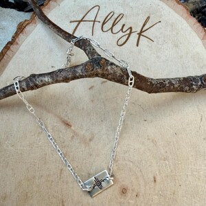 K&ouml;nnte beinhalten: Eine silberne Halskette mit einem rechteckigen Anh&auml;nger mit einem Kreuzdesign. Die Halskette wird auf einer Holzoberfl&auml;che mit dem Namen "Allyk" in Schreibschrift pr&auml;sentiert.