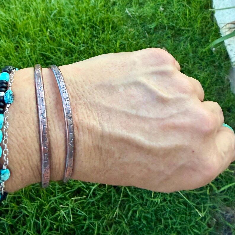 May include: Two thin, metallic bangles featuring a repeating geometric pattern of small squares and lines. Beside them is a beaded bracelet with round turquoise stones and black beads strung on a silver-coloured chain, creating a layered look.