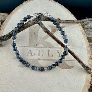 May include: A beaded bracelet featuring round, dark gray, mottled stone beads interspersed with small, ribbed, silver-toned metal spacers. The bracelet is secured with a silver-toned metal lobster claw clasp and is arranged in a circular shape.