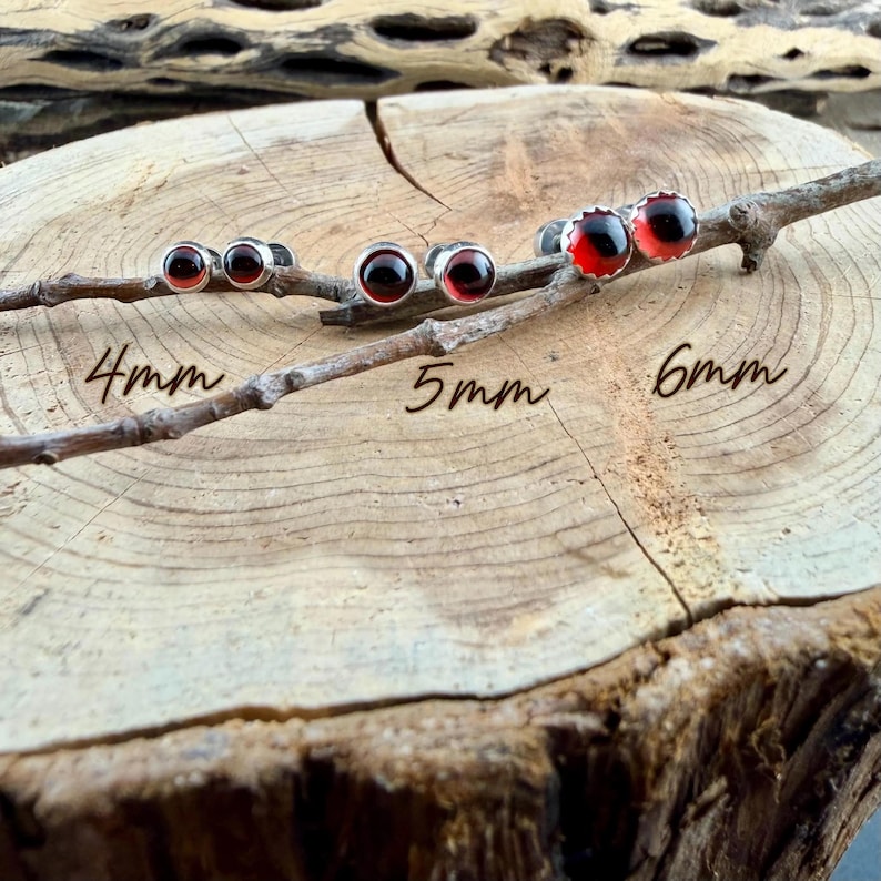 May include: Three pairs of round stud earrings featuring dark red stones set in silver-toned metal. The earrings are labeled with sizes 4mm, 5mm, and 6mm, displayed on a wooden branch. Each pair consists of two circular studs with a smooth finish.