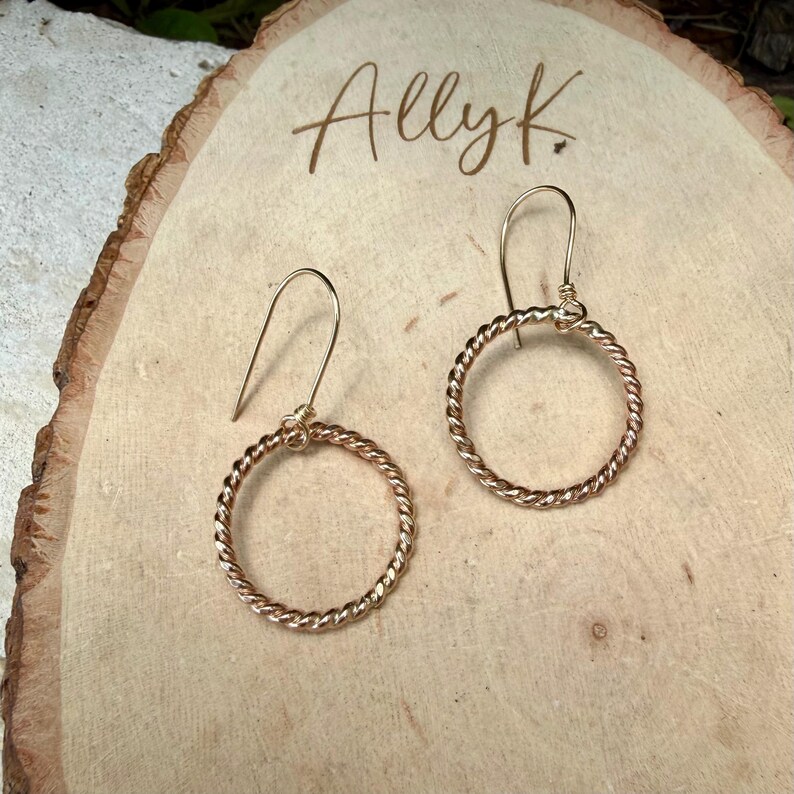 May include: A pair of gold-toned earrings featuring a circular, twisted rope design suspended from simple curved ear wires. The earrings are displayed on a light-colored wooden slice with the text Ally K. engraved in a cursive script at the top.