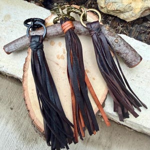 May include: Three leather tassel keychains in black, brown, and tan. Each keychain features a metal ring and a knotted leather top. The tassels are made of long, thin strips of leather, creating a flowing effect. The keychains are displayed on a wooden surface.
