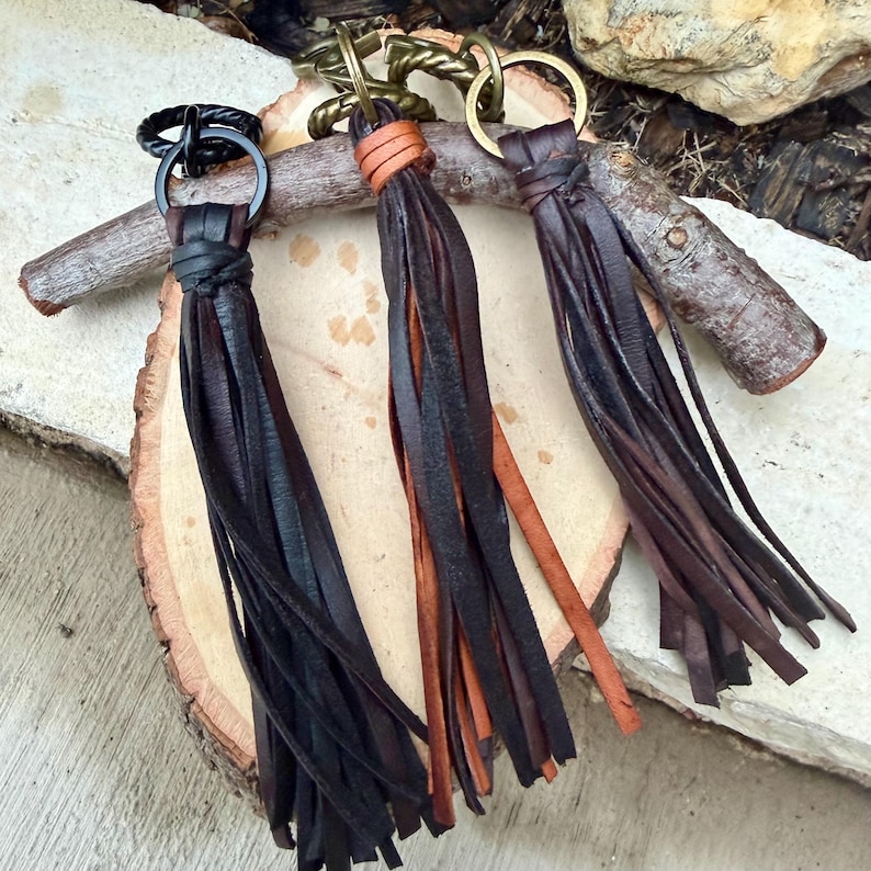 May include: Three leather tassel keychains in black, brown, and tan. Each keychain features a metal ring and a knotted leather top. The tassels are made of long, thin strips of leather, creating a flowing effect. The keychains are displayed on a wooden surface.