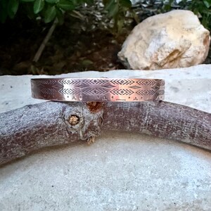 May include: A copper-toned metal cuff bracelet featuring a repeating geometric pattern of etched diamonds, chevrons, and horizontal lines. The band has a smooth, polished finish and a consistent width, showcasing intricate, precise engravings.