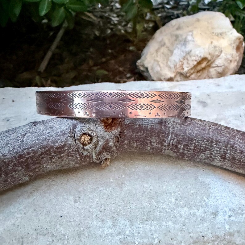 May include: A copper-toned metal cuff bracelet featuring a repeating geometric pattern of etched diamonds, chevrons, and horizontal lines. The band has a smooth, polished finish and a consistent width, showcasing intricate, precise engravings.
