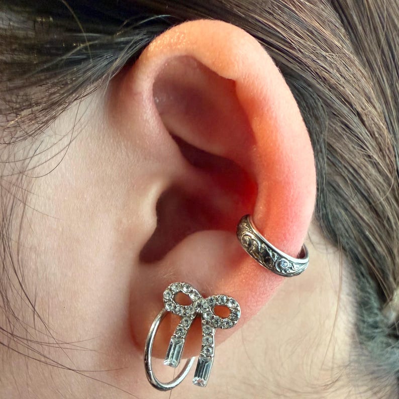 May include: Close-up of an ear with two pieces of jewelry. A silver ear cuff with an engraved design is on the upper ear. A bow-shaped earring with clear crystals and a hoop design is in the earlobe.