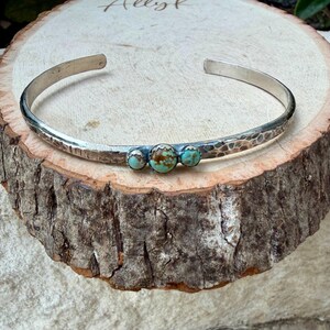 May include: A silver cuff bracelet with a hammered texture and three turquoise and brown stones. The bracelet is displayed on a rustic wooden surface, showcasing its handcrafted design. The open-ended design allows for easy wear.
