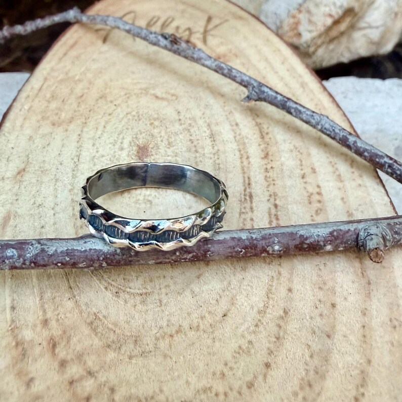 May include: A silver-toned metal ring featuring a textured, wavy band design with dark, recessed detailing between the raised, polished edges. The ring is positioned horizontally, resting against a thin, brown, natural wooden twig.