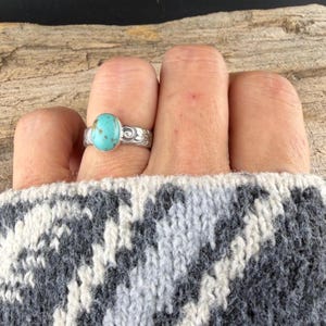 May include: A silver ring with an oval, light blue turquoise stone. The ring band features a detailed floral design. The ring is worn on a finger, with a gray and white patterned sweater in the background.