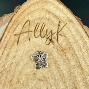 May include: A small, silver-colored butterfly charm with intricate, swirling details. The charm is set against a natural wood slice with the handwritten text "Allyk" carved into the wood.