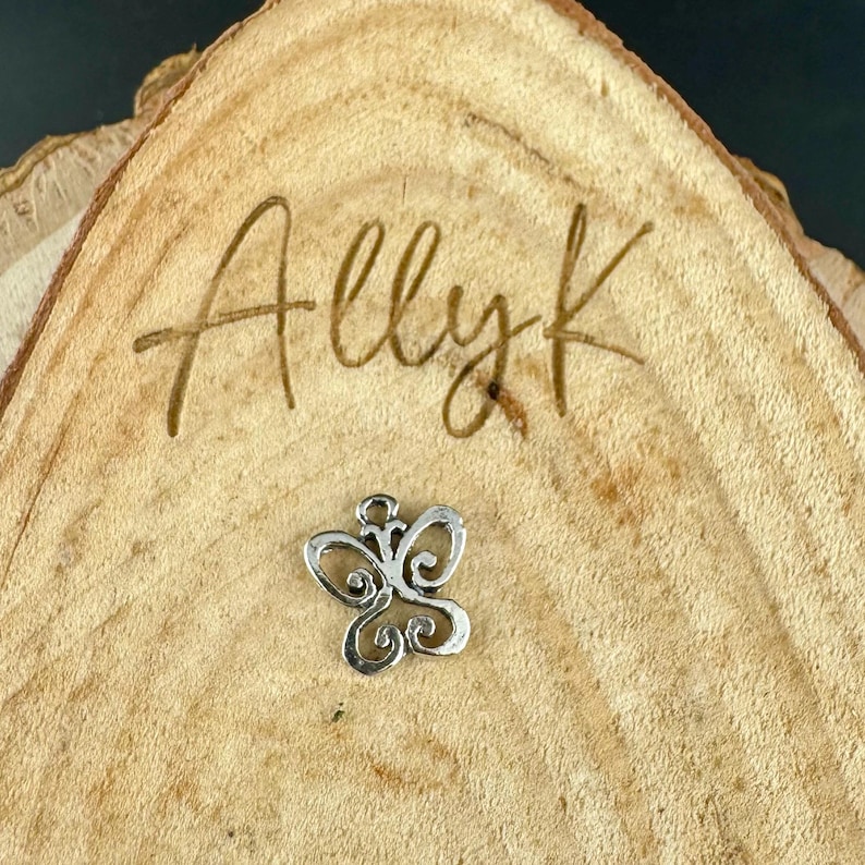 May include: A small, silver-colored butterfly charm with intricate, swirling details. The charm is set against a natural wood slice with the handwritten text "Allyk" carved into the wood.