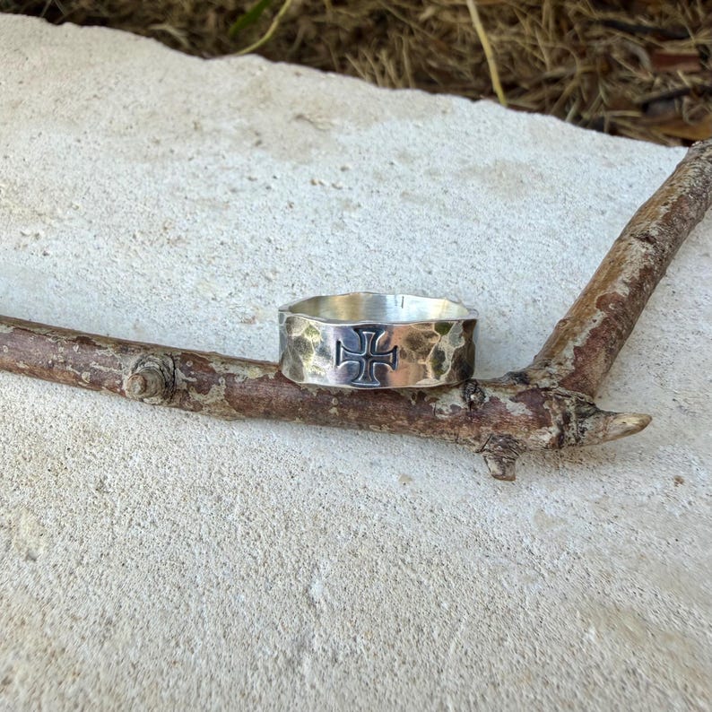 May include: A silver-toned metal ring featuring a hammered, textured surface and a centered cross emblem. The band has a wide, flat profile with a polished finish, showcasing a distinct cross symbol engraved directly into the center of the metal.