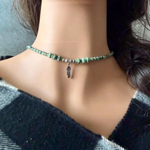 May include: A turquoise beaded choker necklace with a silver feather pendant. The necklace features small, irregular turquoise beads and a delicate silver feather charm. The necklace is worn around the neck.