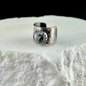 May include: A silver-toned ring with a hammered texture and a round, speckled stone. The stone is set in a scalloped bezel, and the ring has an open design. The ring is displayed on a white, textured surface.