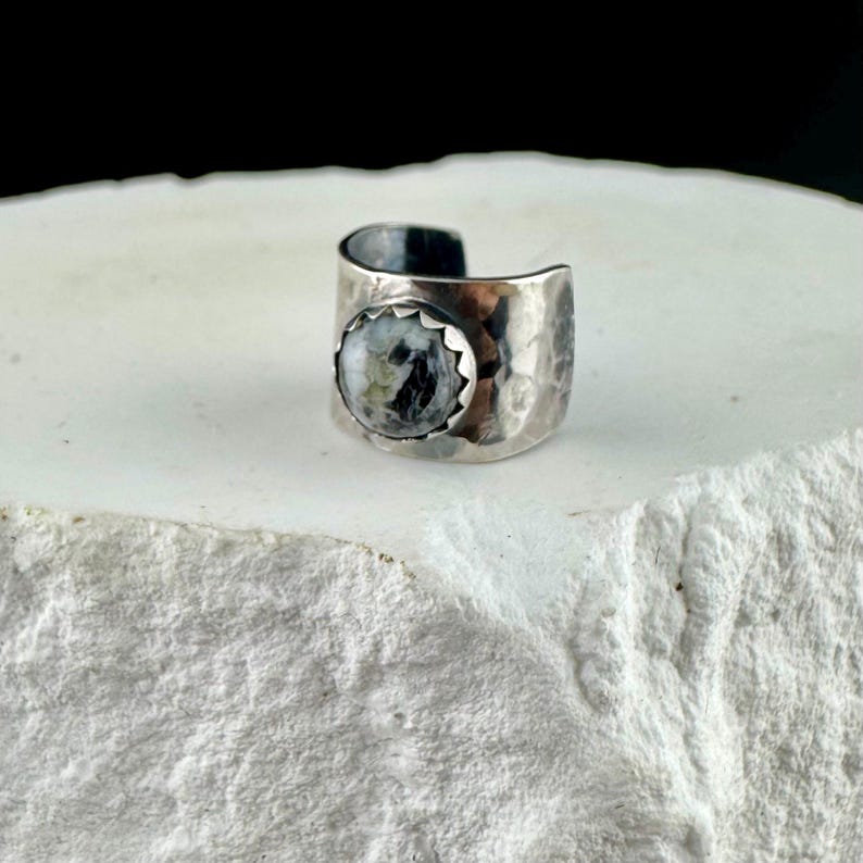 May include: A silver-toned ring with a hammered texture and a round, speckled stone. The stone is set in a scalloped bezel, and the ring has an open design. The ring is displayed on a white, textured surface.