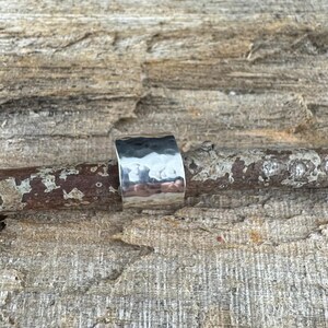May include: A wide, hammered silver ring with a textured surface. The ring is a solid band with a slightly curved shape, reflecting light. The ring is displayed on a piece of weathered wood, showcasing its metallic sheen.