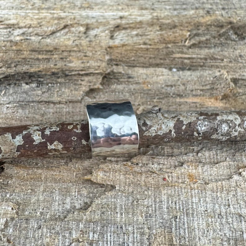 May include: A wide, hammered silver ring with a textured surface. The ring is a solid band with a slightly curved shape, reflecting light. The ring is displayed on a piece of weathered wood, showcasing its metallic sheen.