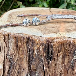 May include: A pair of circular silver stud earrings, each featuring an embossed cross design. The earrings are resting on a thin, light brown twig that is positioned horizontally across the top of a textured, light brown wooden tree stump.