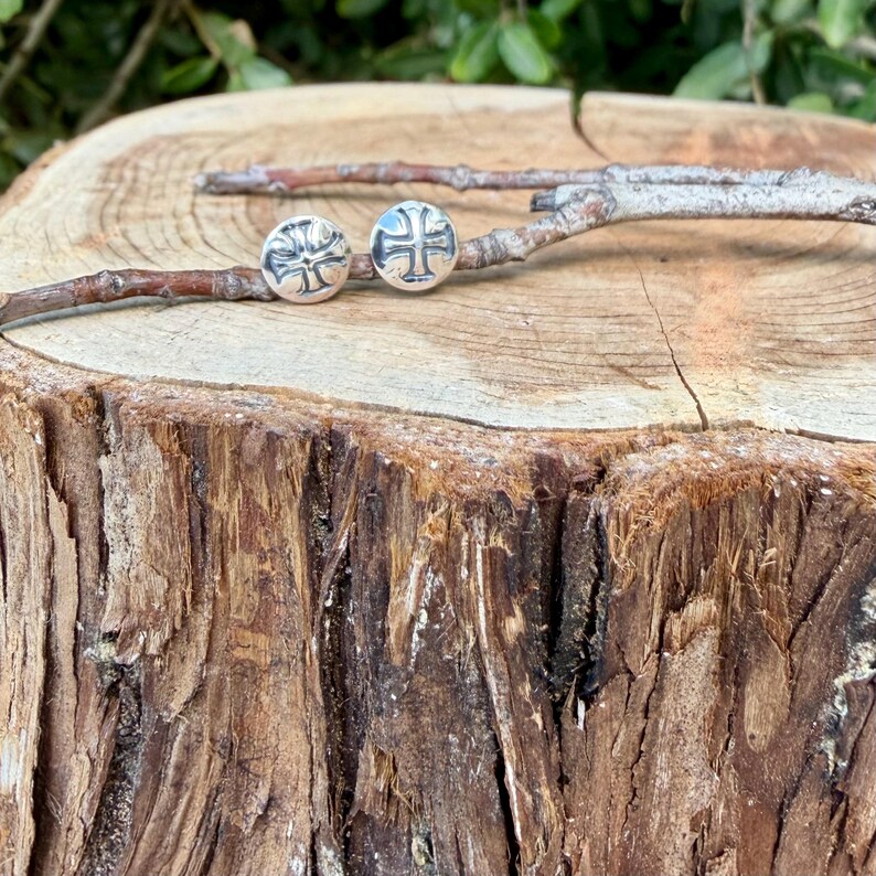 May include: A pair of circular silver stud earrings, each featuring an embossed cross design. The earrings are resting on a thin, light brown twig that is positioned horizontally across the top of a textured, light brown wooden tree stump.