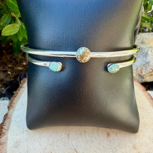 May include: A silver-toned cuff bracelet with three turquoise and brown stones. The bracelet has a simple, open design with a central round stone and two oval stones on the ends. The bracelet is displayed on a black surface.